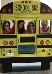 The image features a group of four women standing together, posing for a picture in front of a yellow school bus, they are all smiling.